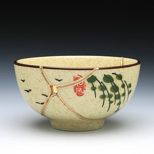 May include: A beige ceramic bowl with a speckled finish, featuring gold-colored kintsugi repairs. The bowl is decorated with black birds, green foliage, and a red emblem. The rim is brown.