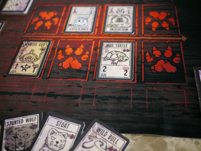May include: A game board with cards featuring animal illustrations and names. The cards are laid out on a red and black game board with paw print symbols. The cards include "Mantis God", "Mud Turtle", "Stunt Wolf", "Stoat", and "Wild Bull".