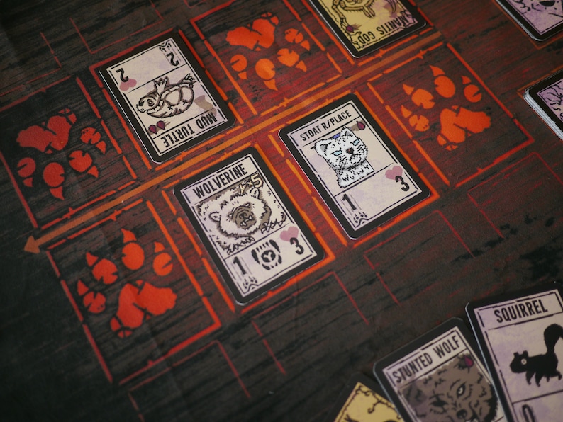 May include: A game board with cards featuring different animals, including a wolverine, a stoat, a mud turtle, a squirrel, and a stunted wolf. The cards have illustrations of the animals and numbers on them. The board has paw prints and the text "Stoat R/Place" and "Mantis Godt".