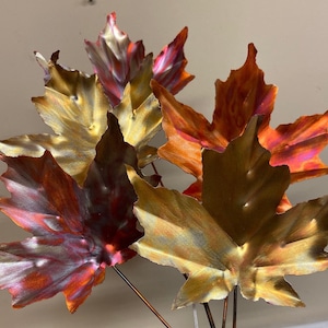 May include: A collection of metal maple leaves in autumnal hues of gold, orange, and burgundy. The leaves are mounted on slender metal stems, forming a decorative arrangement. The leaves have a textured finish and are presented in a natural, organic design.