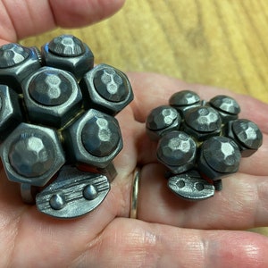 May include: Two metal sculptures of a cluster of hexagonal nuts, one larger than the other. The sculptures are made of silver-coloured metal and have a textured surface.