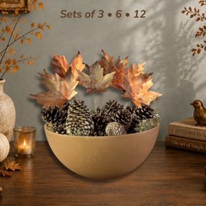 May include: A smooth, matte tan bowl filled with a decorative arrangement of brown pinecones and artificial maple leaves in shades of orange and gold. The text "Sets of 3 • 6 • 12" is displayed above the bowl, which sits on a dark wooden surface.