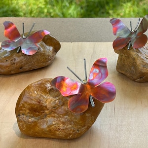 May include: Three decorative metal butterflies with colorful wings are perched on smooth, brown stones. The butterfly wings display shades of pink, orange, and silver. The butterflies are attached to the stones with metal rods.