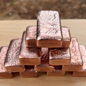 Peut inclure: Une pile de dix barres de cuivre rectangulaires disposées en pyramide. Chaque barre présente une finition métallique brun rougeâtre avec une surface texturée cristalline sur le dessus et des bords arrondis et lisses sur les côtés.