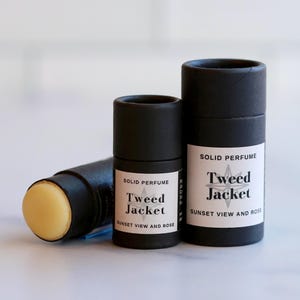 May include: Three black cylindrical containers of solid perfume. One is lying on its side, revealing a yellow balm. The other two are upright, with labels that read "Tweed Jacket, Sunset View and Rose."