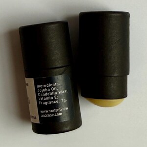 May include: Two black cylindrical tubes with a white label. The label lists ingredients: Jojoba Oil, Candelilla Wax, Vitamin E, and Fragrance. The website address www.sunsetviewandrose.com is also visible. One tube has a yellow balm exposed.