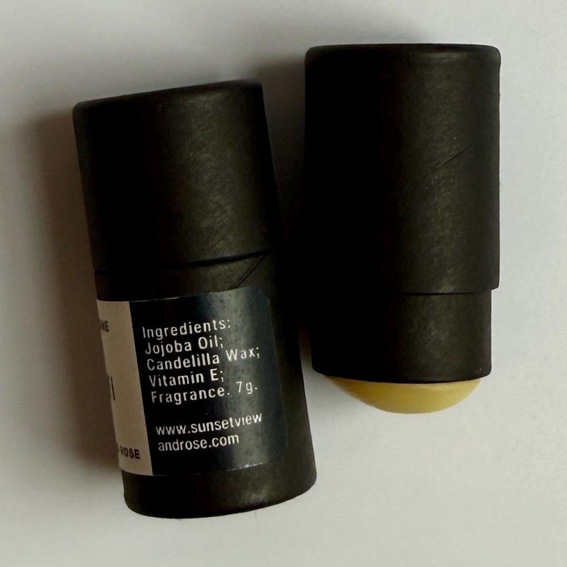 May include: Two black cylindrical tubes with a white label. The label lists ingredients: Jojoba Oil, Candelilla Wax, Vitamin E, and Fragrance. The website address www.sunsetviewandrose.com is also visible. One tube has a yellow balm exposed.