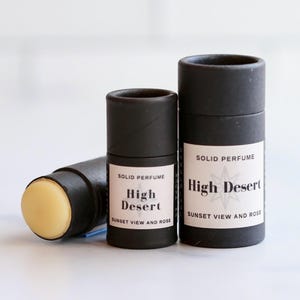 May include: Three black cylindrical containers of solid perfume. One is lying on its side, revealing the solid perfume. The labels read "High Desert, Sunset View and Rose". The other two are upright, with the same label design.