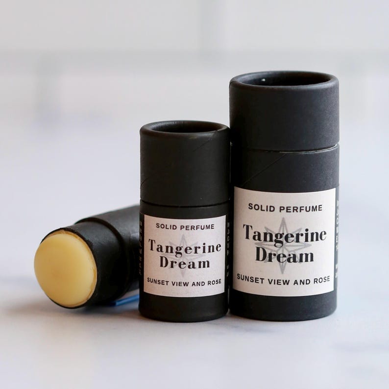 May include: Three black solid perfume containers. Two are upright, and one is on its side, revealing the solid perfume. The labels read "Tangerine Dream, Sunset View and Rose."