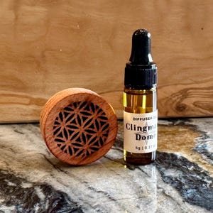 May include: A small wooden diffuser with a geometric pattern and a glass bottle of essential oil labeled "Diffuser Oil Clingman Dome 5g | 0.17 oz".