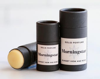 Pode incluir: Três recipientes cilíndricos pretos de perfume sólido. Dois estão em pé e um está de lado, revelando o perfume amarelo claro. Os rótulos dizem "Morningstar, Sunset View and Rose."