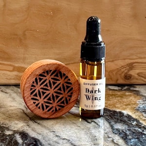 May include: A small brown wooden diffuser with a geometric design and a glass bottle of diffuser oil labeled "Dark Wing" with a 5g (0.17 oz) size.