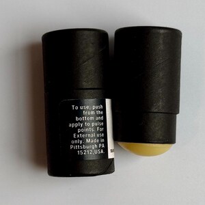 May include: Two black cylindrical containers, one with a visible yellow balm applicator. The containers have a label with instructions: "To use, push from the bottom and apply to pulse points. For External use only. Made in Pittsburgh PA 15212, USA."