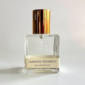 Puede incluir: Un frasco de perfume cuadrado y transparente con una tapa dorada. La etiqueta dice "CASHMERE PATCHOULI Eau de Parfum". El frasco tiene un pulverizador y está sobre un fondo blanco.