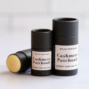 May include: Three black solid perfume containers. One is open, revealing a light yellow balm. The labels read "Cashmere Patchouli, Sunset View and Rose". The containers are cylindrical and made of cardboard.