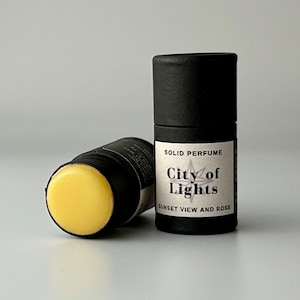 May include: Two black cylindrical solid perfume containers. One is lying on its side, revealing a yellow balm. The other stands upright, displaying a label that reads "City of Lights, Sunset View and Rose."