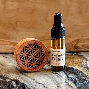May include: A wooden essential oil diffuser with a geometric design next to a small glass bottle of Lavender Fields diffuser oil. The bottle has a black dropper top and a label with the text "Lavender Fields" and "5g | 0.17 oz."