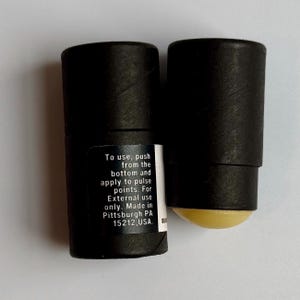 May include: Two black cylindrical containers, one with a visible yellow balm applicator. The containers have a label with instructions: "To use, push from the bottom and apply to pulse points. For External use only. Made in Pittsburgh PA 15212, USA."