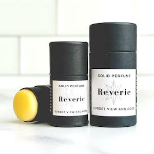 May include: Three black cylindrical solid perfume containers with white labels. The labels read "SOLID PERFUME" and "Reverie Sunset View and Rose." One container is open, revealing a yellow solid perfume stick.