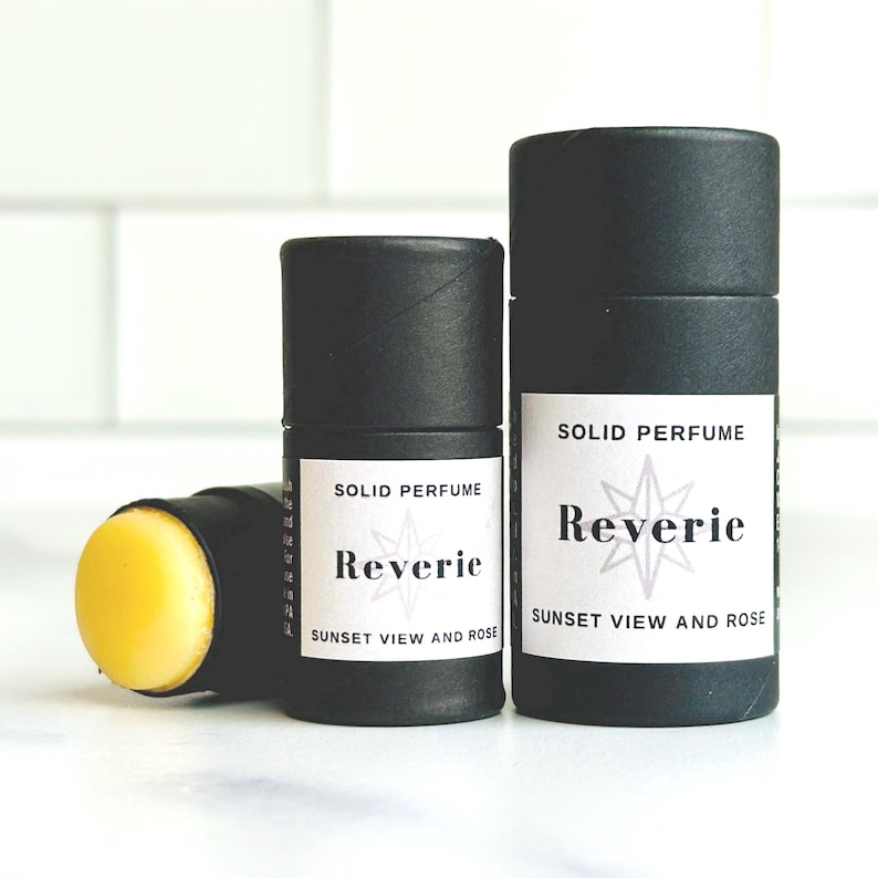 May include: Three black cylindrical solid perfume containers with white labels. The labels read "SOLID PERFUME" and "Reverie Sunset View and Rose." One container is open, revealing a yellow solid perfume stick.