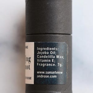 May include: A cylindrical, dark gray product container with a label listing ingredients like Jojoba Oil and Vitamin E. The label also includes the text "Ing not" and the website address www.sunsetviewandrose.com. The product weighs 7g.