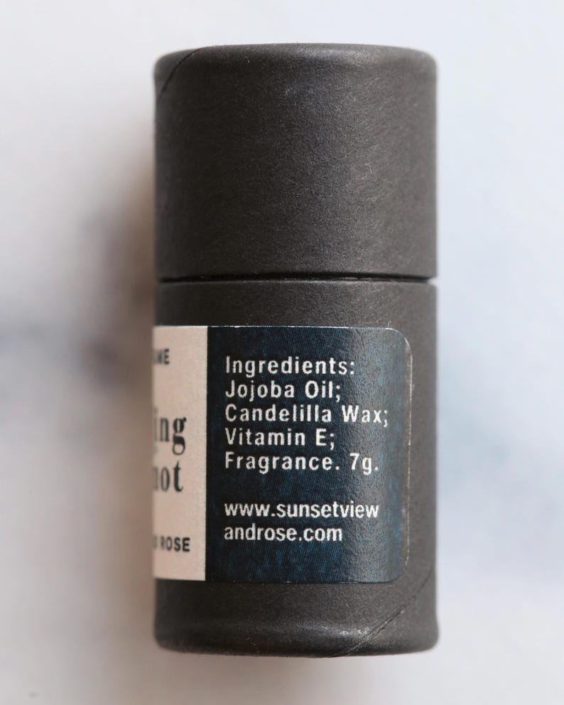May include: A cylindrical, dark gray product container with a label listing ingredients like Jojoba Oil and Vitamin E. The label also includes the text "Ing not" and the website address www.sunsetviewandrose.com. The product weighs 7g.