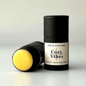 May include: Two black solid perfume sticks. One stick is upright, displaying the label "Cozy Vibes Sunset View and Rose." The other stick is lying down, revealing a yellow, solid perfume. The background is a light gray.