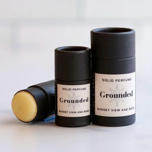 May include: A solid perfume stick and two black cylindrical containers. The stick has a light yellow balm exposed. The containers have white labels with the text "SOLID PERFUME Grounded SUNSET VIEW AND ROSE" and a starburst graphic.