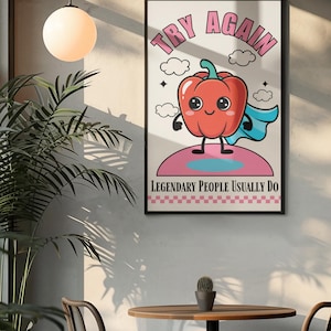 May include: A framed art print with a cartoon red pepper superhero. The text "TRY AGAIN" is at the top, and "LEGENDARY PEOPLE USUALLY DO" is at the bottom. The artwork has a retro, whimsical style, with a pink and blue colour scheme.