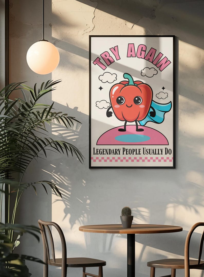 May include: A framed art print with a cartoon red pepper superhero. The text "TRY AGAIN" is at the top, and "LEGENDARY PEOPLE USUALLY DO" is at the bottom. The artwork has a retro, whimsical style, with a pink and blue colour scheme.