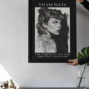 May include: Black framed art print featuring a grayscale portrait of Sylvia Plath. The print includes the author's name at the top and a quote at the bottom. The artwork is held up against a white wall.