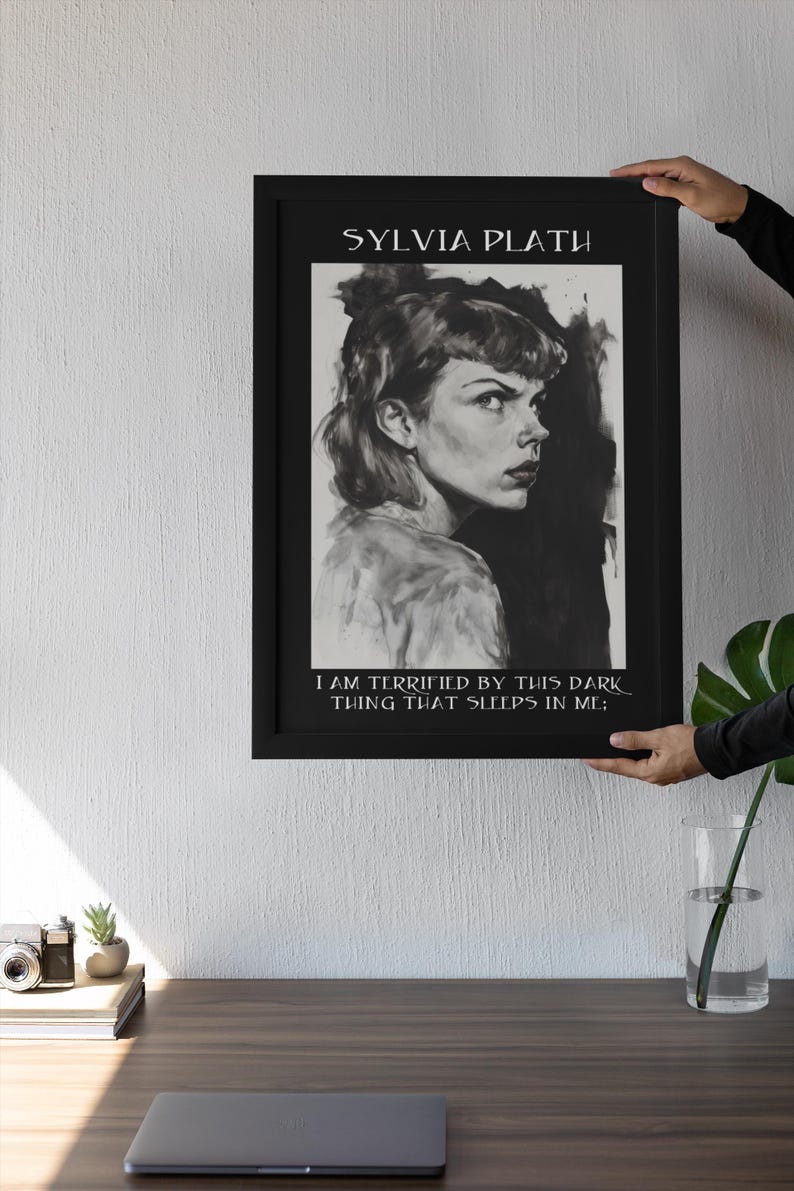 May include: Black framed art print featuring a grayscale portrait of Sylvia Plath. The print includes the author's name at the top and a quote at the bottom. The artwork is held up against a white wall.