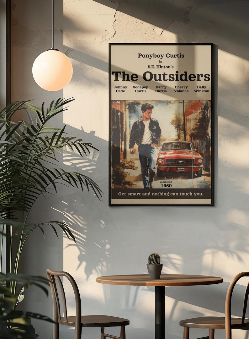 Op de afbeelding: Een rechthoekige wandposter met de titel The Outsiders van S.E. Hinton. Het kunstwerk toont een persoon in een donker jasje en blauwe spijkerbroek die bij een rode auto loopt. De tekst bevat karakternamen, published 1969 en Get smart and nothing can touch you.