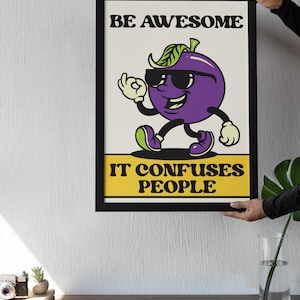 May include: Framed art print with a cartoon plum character wearing sunglasses and giving an 'okay' hand gesture. The text reads 'BE AWESOME' above the plum and 'IT CONFUSES PEOPLE' below. The artwork is in a black frame.