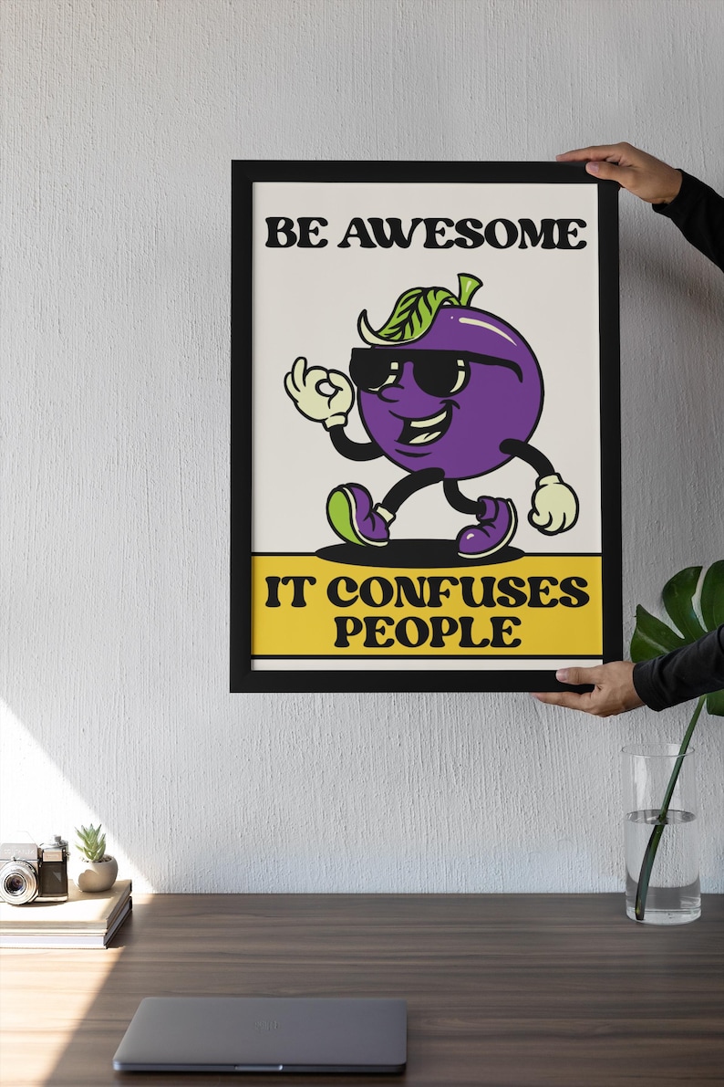 May include: Framed art print with a cartoon plum character wearing sunglasses and giving an 'okay' hand gesture. The text reads 'BE AWESOME' above the plum and 'IT CONFUSES PEOPLE' below. The artwork is in a black frame.