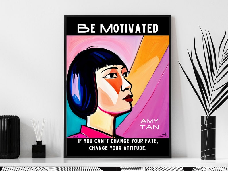 May include: A framed art print with the words "BE MOTIVATED" at the top. The artwork features a stylized portrait of a woman with black hair and a pink and orange background. The text "AMY TAN" and a motivational quote are also included.