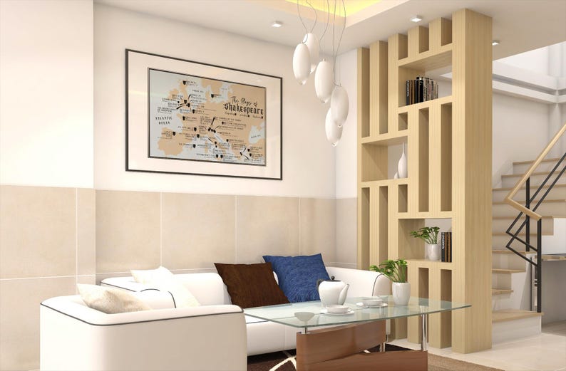 May include: A white sectional sofa with brown and blue throw pillows sits in a living room with a wooden bookshelf and a framed map of Shakespeare's plays on the wall.