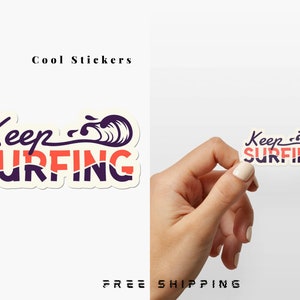 May include: A white sticker with the text "Keep Surfing" in purple and orange lettering. A stylized wave graphic is behind the text. The sticker is being held by a hand.