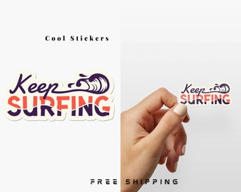 May include: A white sticker with the text "Keep Surfing" in purple and orange lettering. A stylized wave graphic is behind the text. The sticker is being held by a hand.
