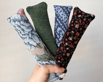 May include: Four catnip toys in different patterns. The toys are made of fabric and are shaped like long, thin pillows. The toys are held in a hand.