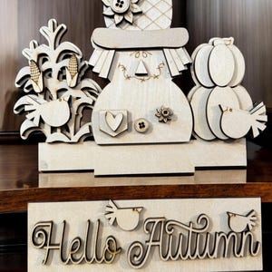 May include: Wooden fall-themed decoration featuring a scarecrow, pumpkins, and corn stalks. The scarecrow has a hat with a flower and button details. A sign below reads "Hello Autumn" with bird accents. The decoration is made of light-colored wood.