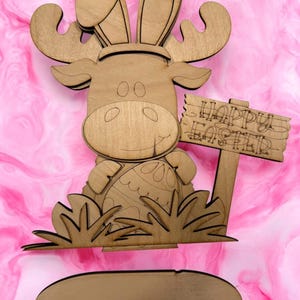 May include: Wooden cutout of a moose with bunny ears holding an Easter egg. The moose is standing in grass and holding a sign that says "Happy Easter".