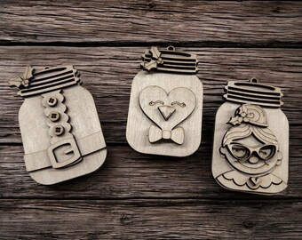 May include: Three wooden mason jar ornaments with festive designs. One features a Santa suit, another a heart-shaped penguin, and the last a woman with glasses. Each has a decorative lid and a hole for hanging.