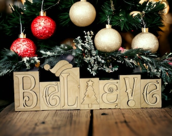 May include: Wooden block letters spelling "Believe" with a Santa hat on the letter "L" and a Christmas tree on the letter "E". The letters are on a wooden surface in front of a Christmas tree decorated with red and gold ornaments.