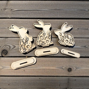 May include: Three wooden Easter bunny decorations with floral cutouts and separate stands. The bunnies are light brown and have intricate details. The stands are oval-shaped with a slot for the bunny to stand in.