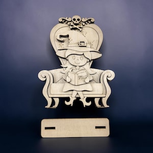 May include: A light-colored wooden cutout featuring a character wearing a witch hat and sitting on a chair. The character holds an open book with the word SPELLS printed on it. A skull and crossbones design sits at the top of the chair back.