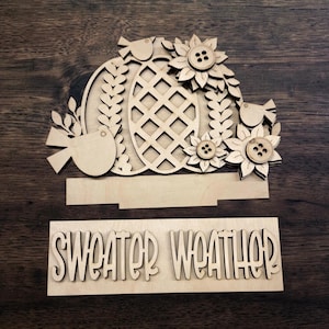 May include: Laser-cut wooden fall-themed decoration featuring a pumpkin, sunflowers, and birds. The pumpkin has a lattice design. The phrase "SWEATER WEATHER" is also included in the design. The decoration is made of unfinished wood.