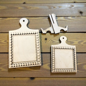 May include: Three unfinished wooden cutting boards with a decorative scalloped border and a handle. One board is larger than the other. A wooden stand is also included. The items are displayed against a wooden background.