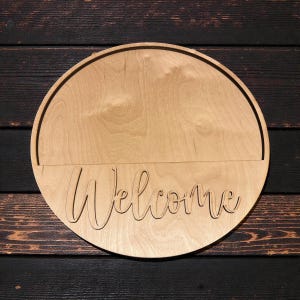 May include: A round wooden sign with the word "Welcome" cut out in a cursive font. The sign is made of light-colored wood and has a dark brown border. The background is a dark wood surface.
