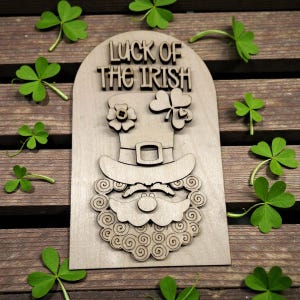 May include: Wooden St. Patrick's Day decoration featuring a leprechaun hat, beard, and the text "Luck of the Irish." The design includes a flower and a shamrock. Green shamrocks are scattered around the wooden decoration on a wooden surface.
